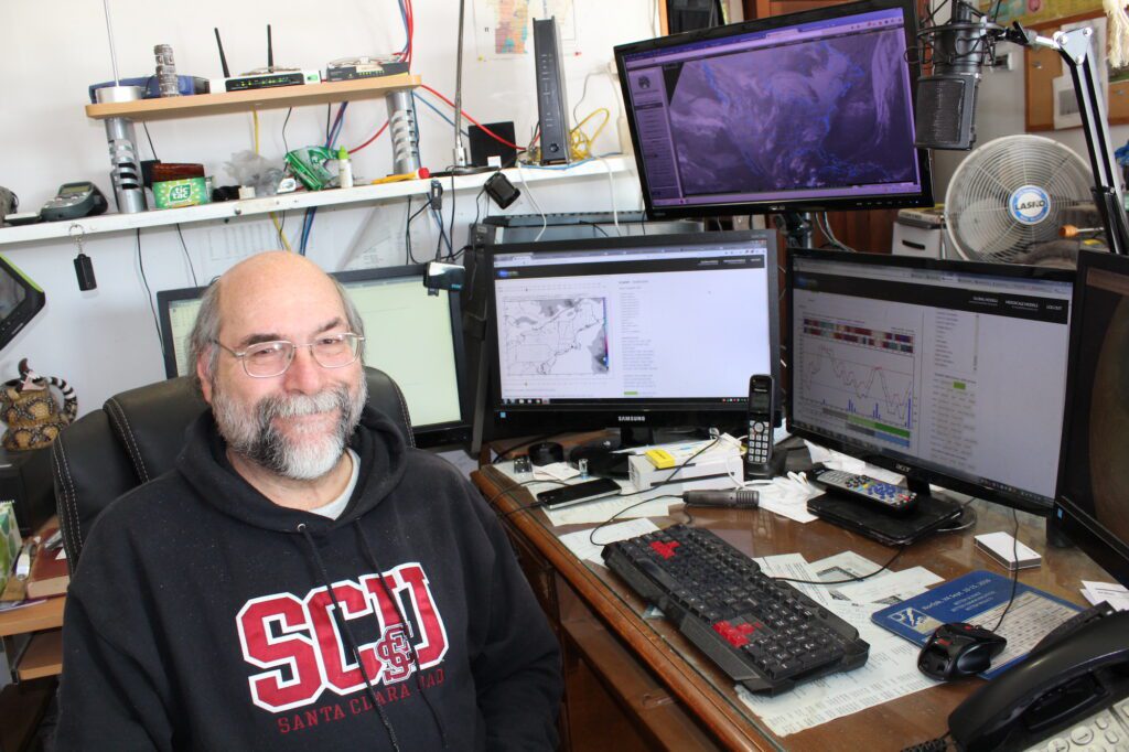 Man with white beard, glasses, light skin, no hair, wearing a black sweatshirt with SCU in red letters, sits in front of three computer monitors at a home office desk